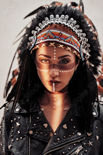 Illuminated with sunlight and confident female wearing American indian traditional headdress, leather jacket and sunglasses while posing in a bright studio and looking serious
