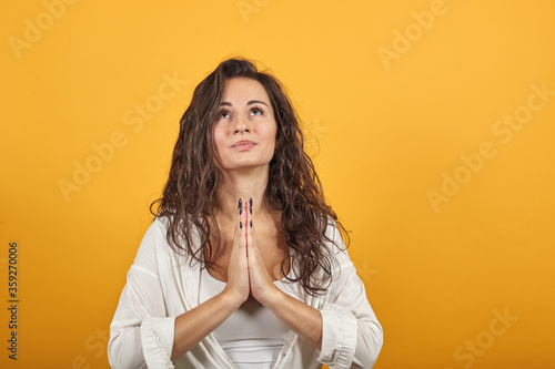 Folded hands together praying asking god for help ask implore wish upwards jesus christ. Young attractive woman, dressed white blouse, with brown eyes, curly hair, yellow background