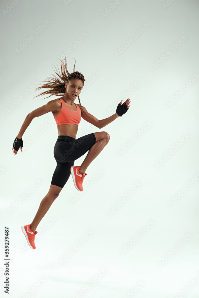 Do it! Full length of young african woman with perfect body in sports clothing jumping in studio against grey background and looking at camera