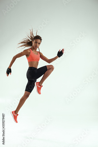 Do it! Full length of young african woman with perfect body in sports clothing jumping in studio against grey background and looking at camera