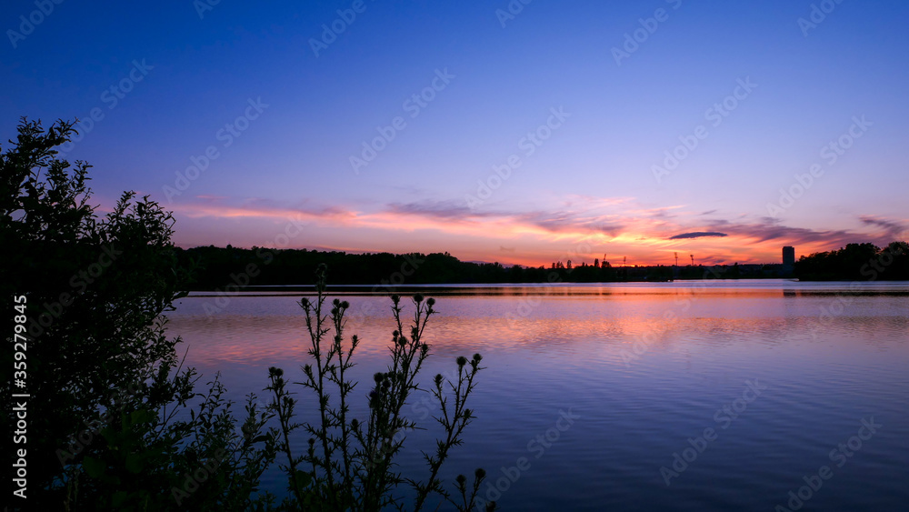 Symmetry of the sky in a lake at sunrise. Clouds reflecting on the water. Holiday landscape by the sea. Quiet relaxing scene with a beautiful colorful sky. Vegetation in the foreground.