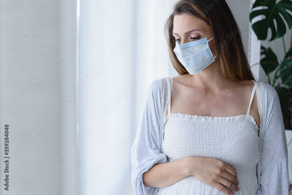 Pregnant woman standing by the big window with face medical mask on. Worries about childbirth during a pandemic. Pregnant woman standing by the big window with face medical mask on. Worries about childbirth during a pandemic.