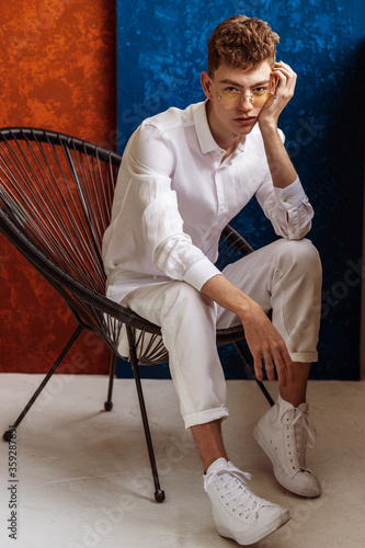 Fashionable young handsome confident man wearing trendy glasses, white linen shirt, trousers, sneakers, posing in colorful interior. Summer fashion conception. Full-length portrait