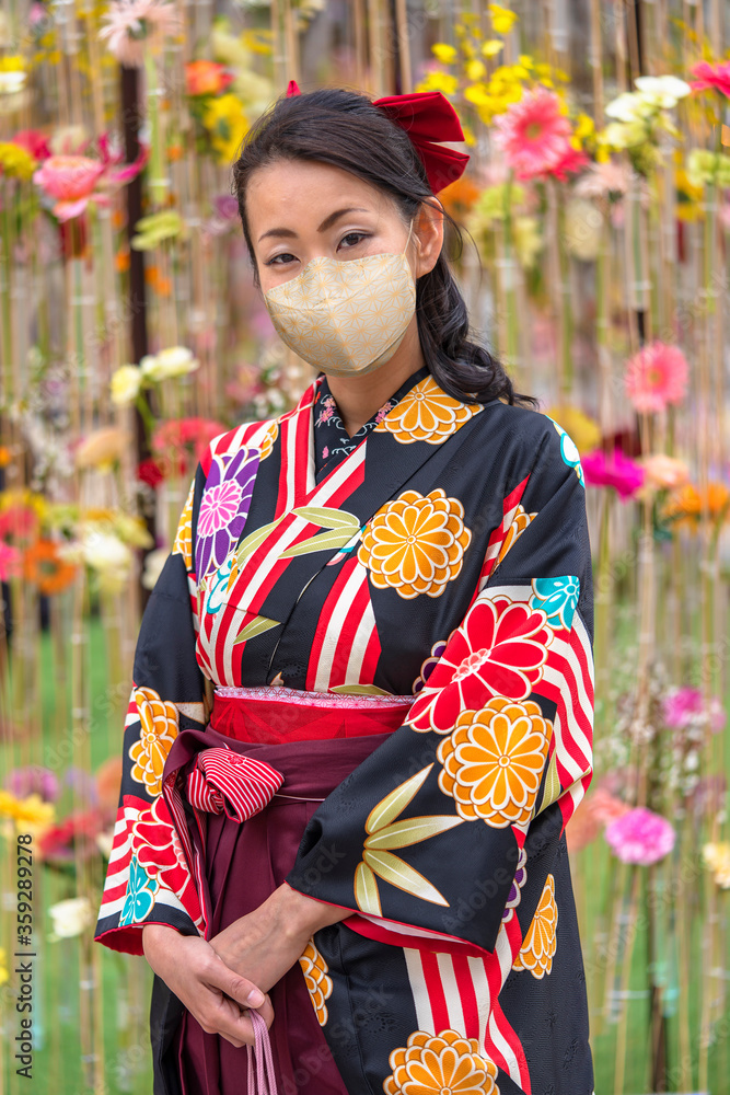Japanese woman wearing a face mask in hakama kimono in front of a wall of flowers