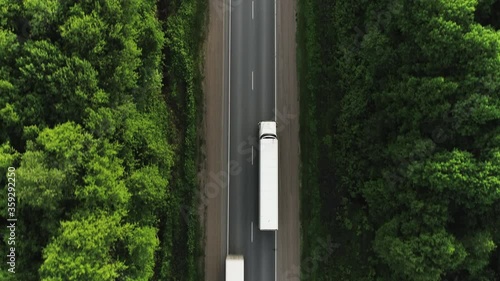 One Semi Truck with white trailer and cab driving / traveling alone on dense flat forest asphalt straight empty road, highway top down view follow vehicle aerial footage / Freeway trucks traffic