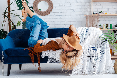 Boho style in wear and interior. Happy smiling woman  laying on blue sofa, posing in cozy room with green plants, white walls, trendy decor. Model hides her face with hat photo