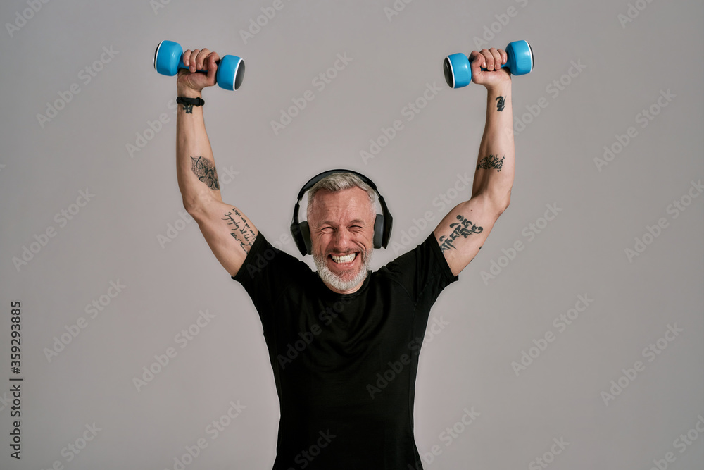 Do it for life. Middle aged muscular man in black t shirt and headphones closing eyes while holding blue dumbbells, posing in studio over grey background Do it for life. Middle aged muscular man in black t shirt and headphones closing eyes while holding blue dumbbells, posing in studio over grey background