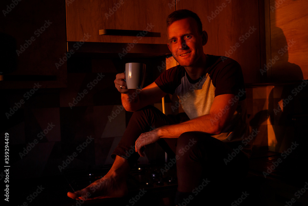 A man sits in the kitchen in the sunset light and drinks coffee