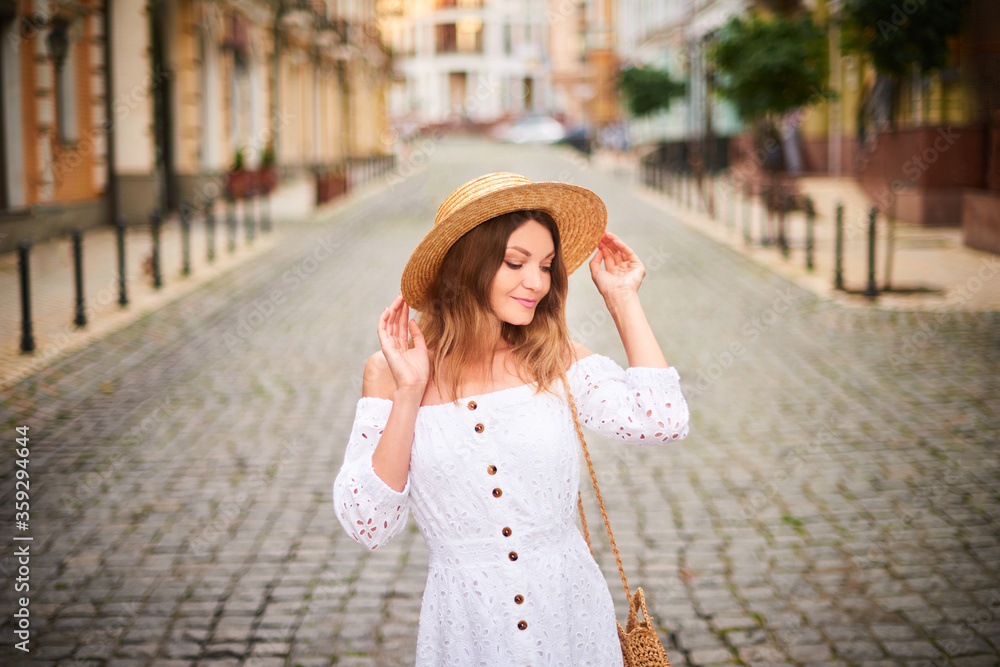 Portrait cheerful tourist woman walk on ancient street in old town. Attractive woman in straw hat, white sundress enjoying solo vacation in Europe. Tourism and travel concept.
