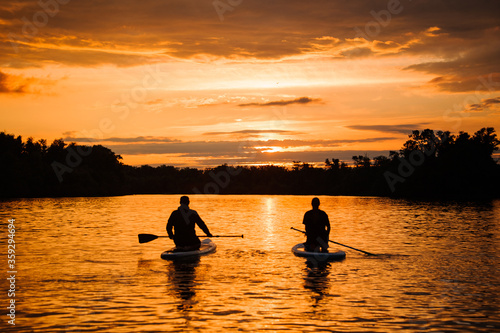 rear view of two sitting people on sup boards which floating on the river at sunset