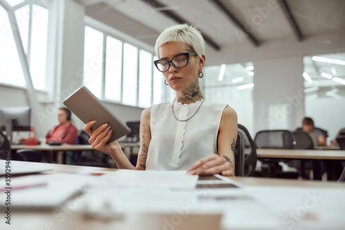 Smart woman. Young and beautiful tattooed business woman wearing glasses working with digital tablet while sitting at her workplace in the office