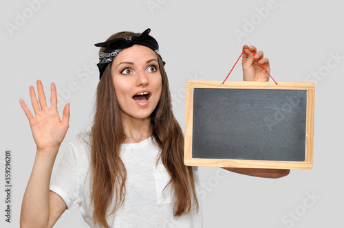 Bild auf Leinwand A young dark-haired woman wearing a white T-shirt and a headband, looking off to the side in surprise and holding an empty chalkboard in a wooden frame
