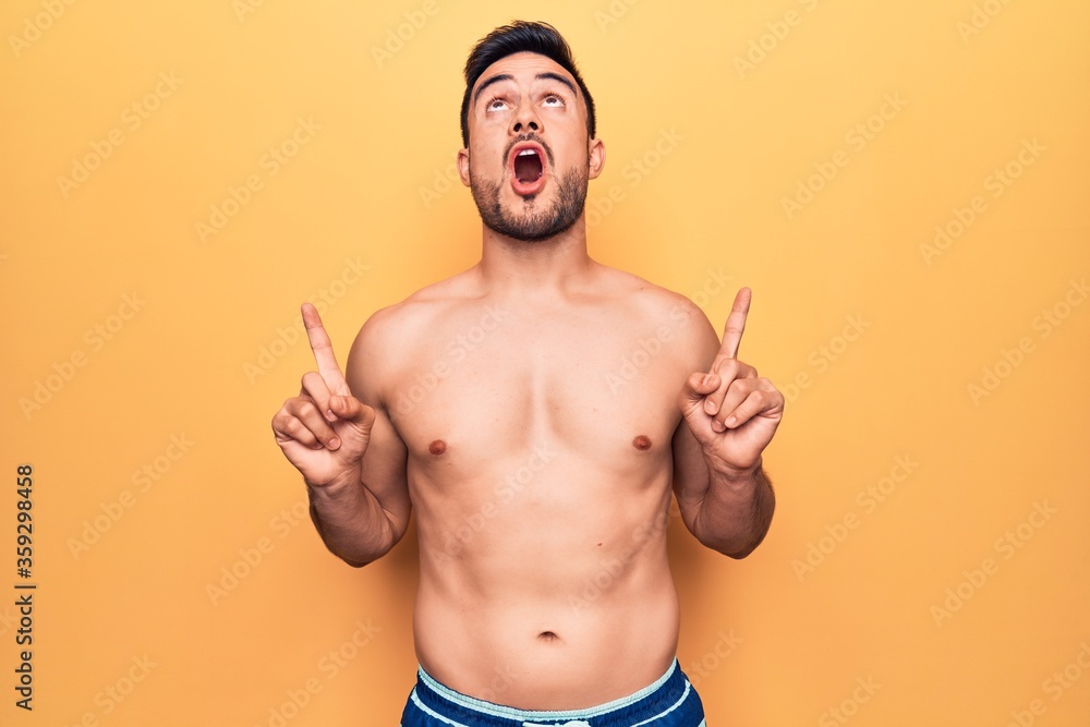 Young handsome man with beard wearing sleeveless t-shirt standing over yellow background amazed and surprised looking up and pointing with fingers and raised arms.