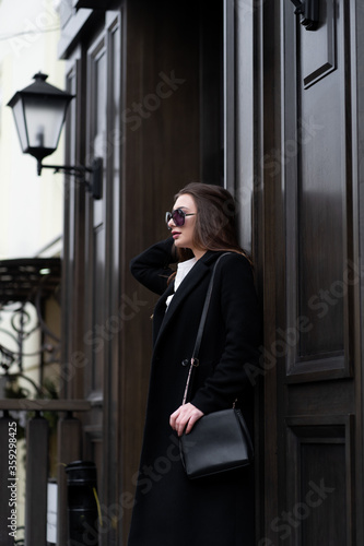 Outdoor photo of brunette lady walking on street background in cold autumn day. Fashion street style portrait. wearing dark casual trousers, white shirt, sunglasses and black coat. Fashion concept.