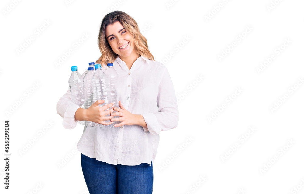 Young caucasian woman holding recycling plastic bottles looking positive and happy standing and smiling with a confident smile showing teeth