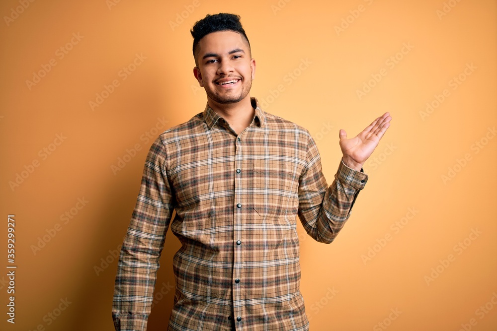 Young handsome man wearing casual shirt standing over isolated yellow background smiling cheerful presenting and pointing with palm of hand looking at the camera. Young handsome man wearing casual shirt standing over isolated yellow background smiling cheerful presenting and pointing with palm of hand looking at the camera.