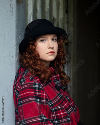 Portrait of a young woman with curly red hair, wearing a red tartan coat, set in a rough urban setting.