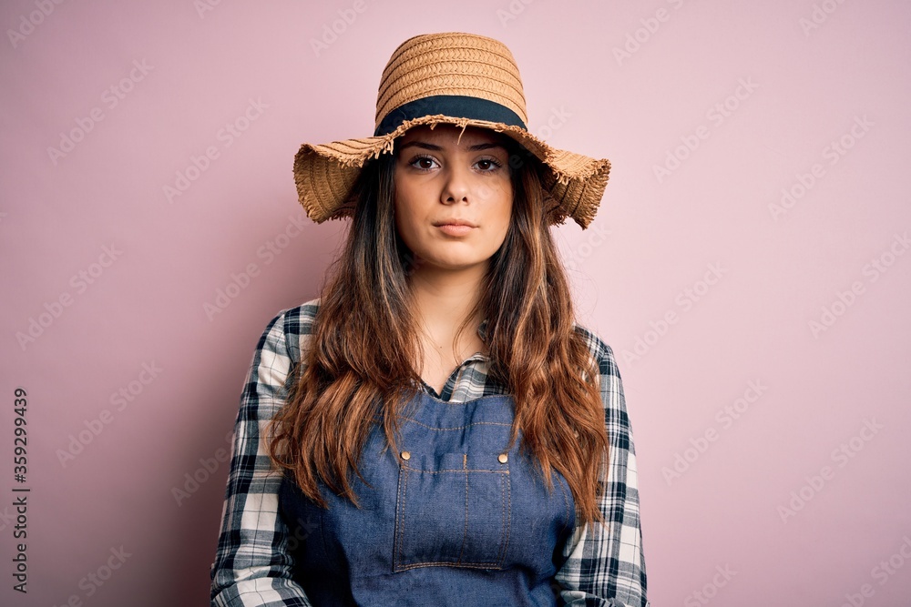 Young beautiful brunette farmer woman wearing apron and hat over pink background with serious expression on face. Simple and natural looking at the camera. Young beautiful brunette farmer woman wearing apron and hat over pink background with serious expression on face. Simple and natural looking at the camera.