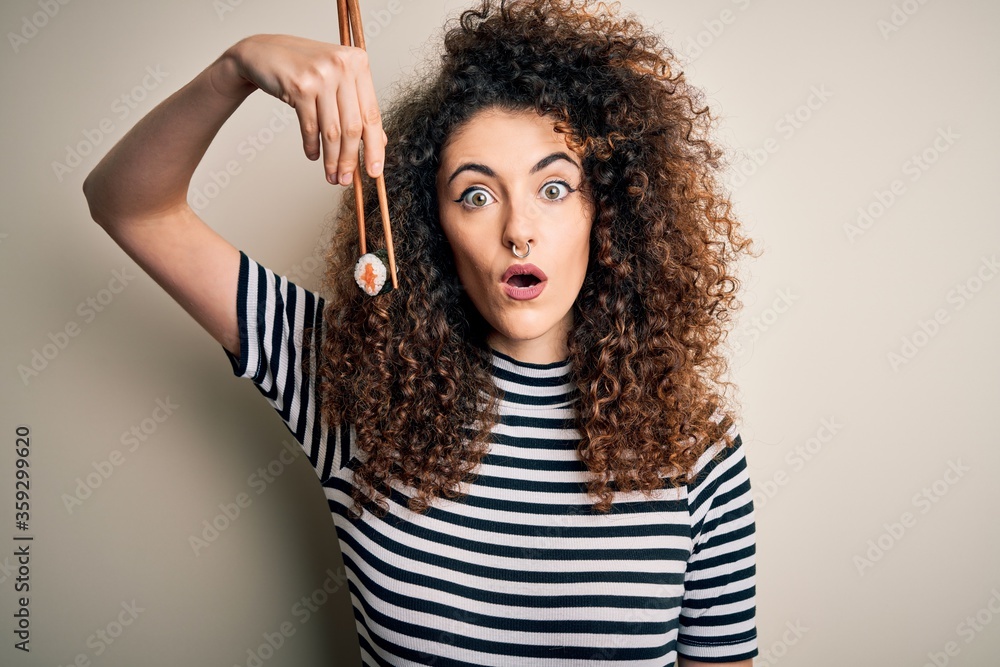 Young beautiful woman with curly hair and piercing eating sushi using wooden chopsticks scared in shock with a surprise face, afraid and excited with fear expression Young beautiful woman with curly hair and piercing eating sushi using wooden chopsticks scared in shock with a surprise face, afraid and excited with fear expression