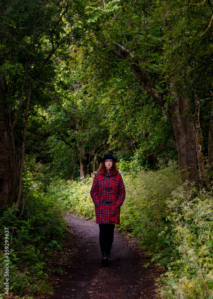 Young woman with red curly hair and wearing a black hat and red tartan coat walking on path in woodland. Young woman with red curly hair and wearing a black hat and red tartan coat walking on path in woodland.