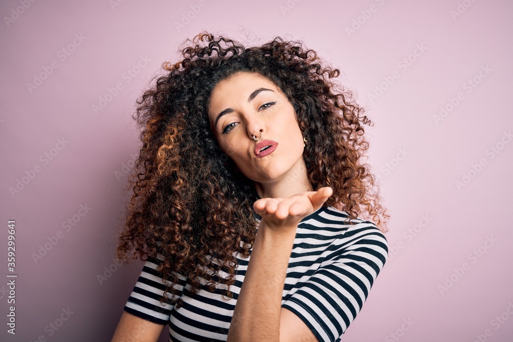 Young beautiful woman with curly hair and piercing wearing casual striped t-shirt looking at the camera blowing a kiss with hand on air being lovely and sexy. Love expression. Young beautiful woman with curly hair and piercing wearing casual striped t-shirt looking at the camera blowing a kiss with hand on air being lovely and sexy. Love expression.