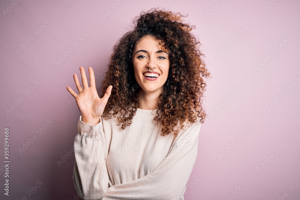 Beautiful woman with curly hair and piercing wearing casual sweater over pink background showing and pointing up with fingers number five while smiling confident and happy. Beautiful woman with curly hair and piercing wearing casual sweater over pink background showing and pointing up with fingers number five while smiling confident and happy.