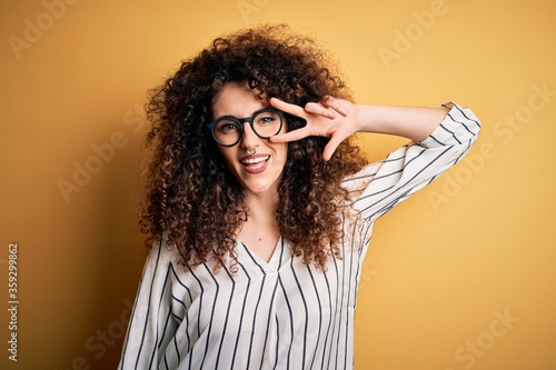 Young beautiful woman with curly hair and piercing wearing striped shirt and glasses Doing peace symbol with fingers over face, smiling cheerful showing victory