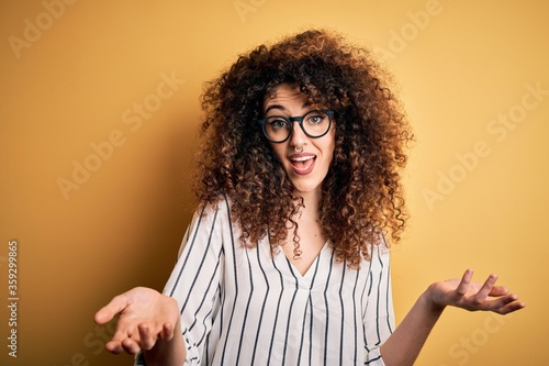 Young beautiful woman with curly hair and piercing wearing striped shirt and glasses smiling cheerful offering hands giving assistance and acceptance.