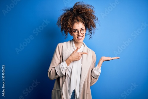 Young beautiful businesswoman with curly hair and piercing wearing jacket and glasses amazed and smiling to the camera while presenting with hand and pointing with finger.