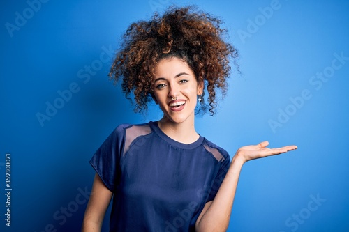 Young beautiful woman with curly hair and piercing wearing casual blue t-shirt smiling cheerful presenting and pointing with palm of hand looking at the camera.