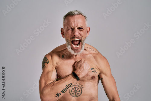 Inner power. Close up of half naked middle aged muscular man shouting at camera, beating his chest while posing in studio over grey background