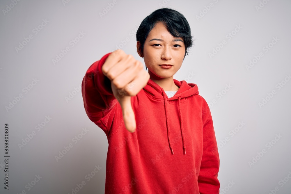 Young beautiful asian girl wearing casual sweatshirt with hoodie over white background looking unhappy and angry showing rejection and negative with thumbs down gesture. Bad expression. Young beautiful asian girl wearing casual sweatshirt with hoodie over white background looking unhappy and angry showing rejection and negative with thumbs down gesture. Bad expression.