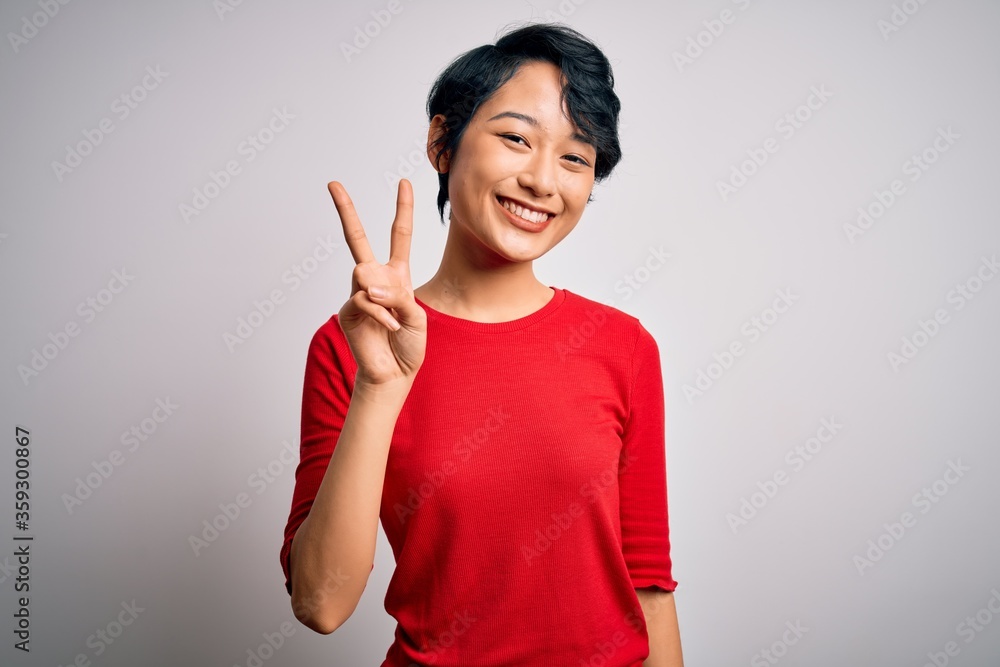 Young beautiful asian girl wearing casual red t-shirt standing over isolated white background smiling looking to the camera showing fingers doing victory sign. Number two. Young beautiful asian girl wearing casual red t-shirt standing over isolated white background smiling looking to the camera showing fingers doing victory sign. Number two.