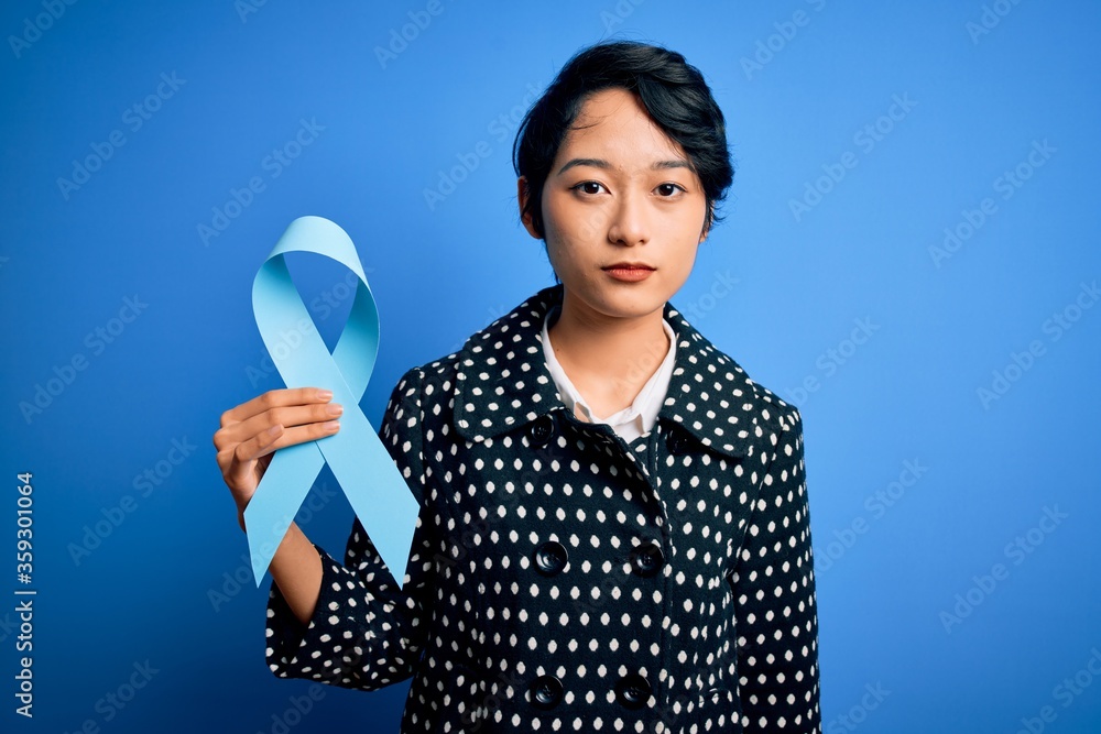 Young beautiful asian girl holding blue cancer ribbon symbol over isolated background with a confident expression on smart face thinking serious Young beautiful asian girl holding blue cancer ribbon symbol over isolated background with a confident expression on smart face thinking serious