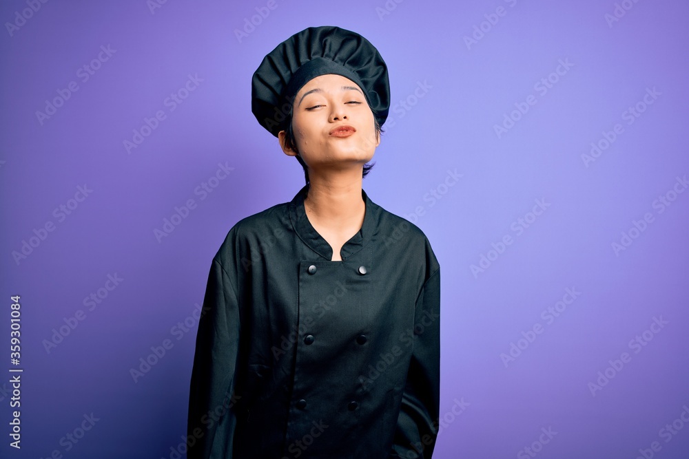 Young beautiful chinese chef woman wearing cooker uniform and hat over purple background looking at the camera blowing a kiss on air being lovely and sexy. Love expression.