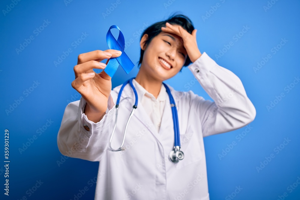 Young beautiful asian doctor girl wearing stethoscope and coat holding blue cancer ribbon stressed with hand on head, shocked with shame and surprise face, angry and frustrated. Fear and upset. Young beautiful asian doctor girl wearing stethoscope and coat holding blue cancer ribbon stressed with hand on head, shocked with shame and surprise face, angry and frustrated. Fear and upset.