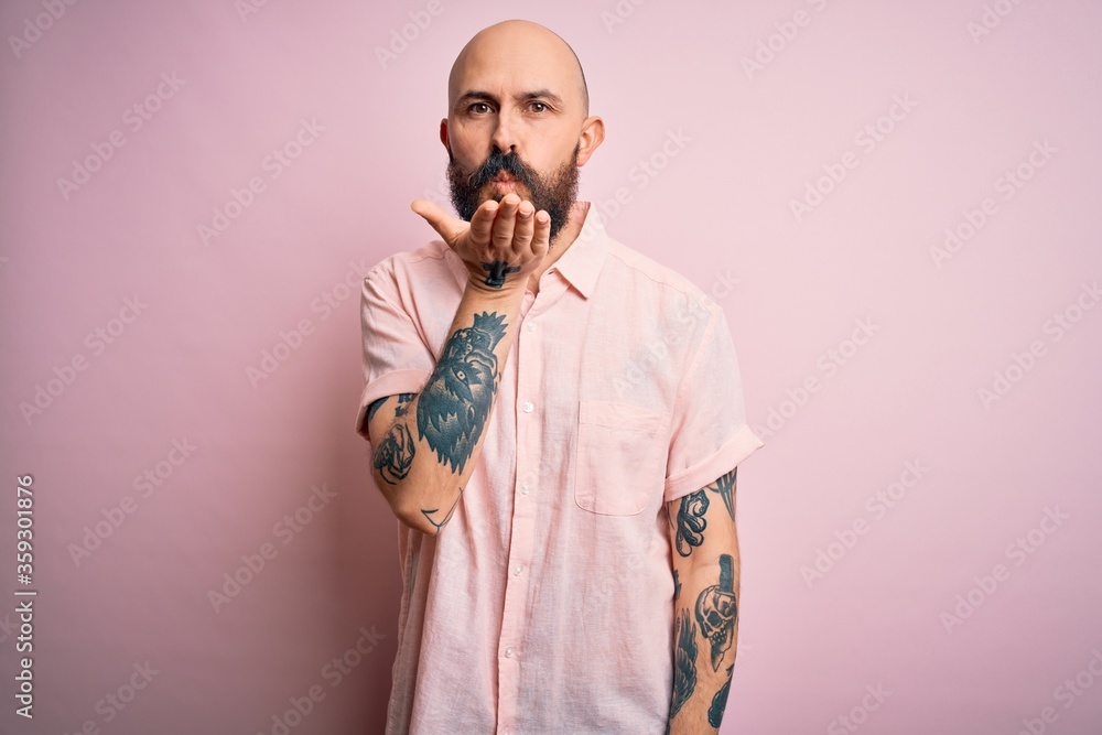 Handsome bald man with beard and tattoo wearing casual shirt over isolated pink background looking at the camera blowing a kiss with hand on air being lovely and sexy. Love expression. Handsome bald man with beard and tattoo wearing casual shirt over isolated pink background looking at the camera blowing a kiss with hand on air being lovely and sexy. Love expression.