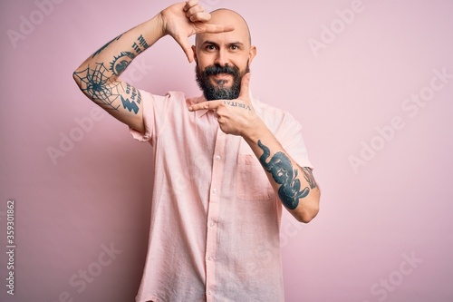 Handsome bald man with beard and tattoo wearing casual shirt over isolated pink background smiling making frame with hands and fingers with happy face. Creativity and photography concept. © Krakenimages.com