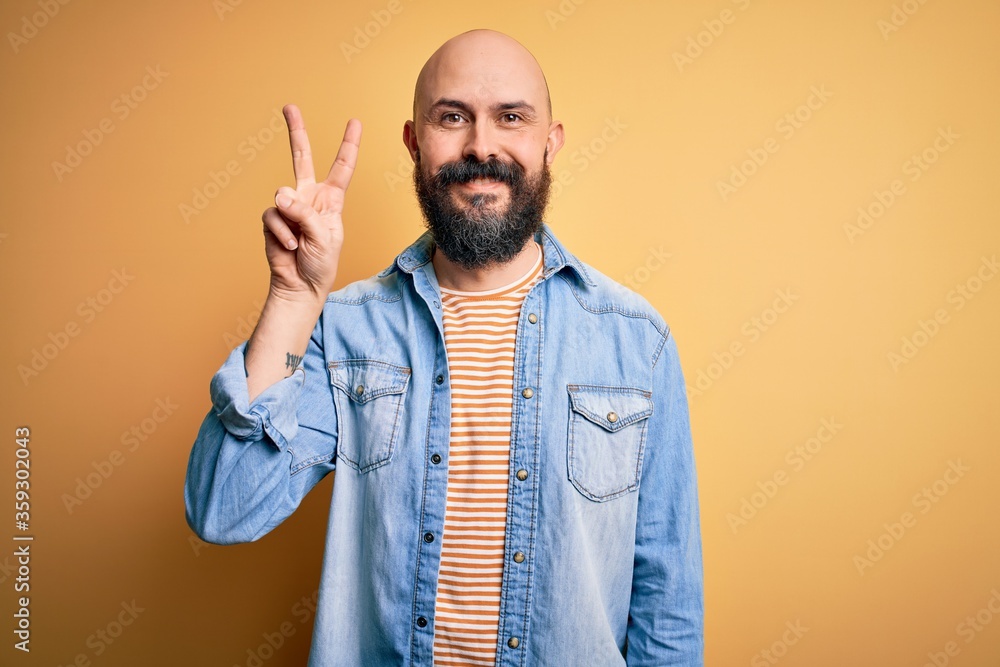Handsome bald man with beard wearing casual denim jacket and striped t-shirt showing and pointing up with fingers number two while smiling confident and happy. Handsome bald man with beard wearing casual denim jacket and striped t-shirt showing and pointing up with fingers number two while smiling confident and happy.