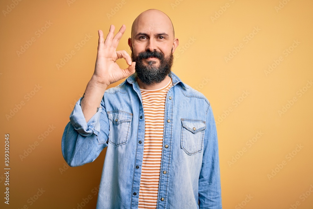 Handsome bald man with beard wearing casual denim jacket and striped t-shirt smiling positive doing ok sign with hand and fingers. Successful expression. Handsome bald man with beard wearing casual denim jacket and striped t-shirt smiling positive doing ok sign with hand and fingers. Successful expression.