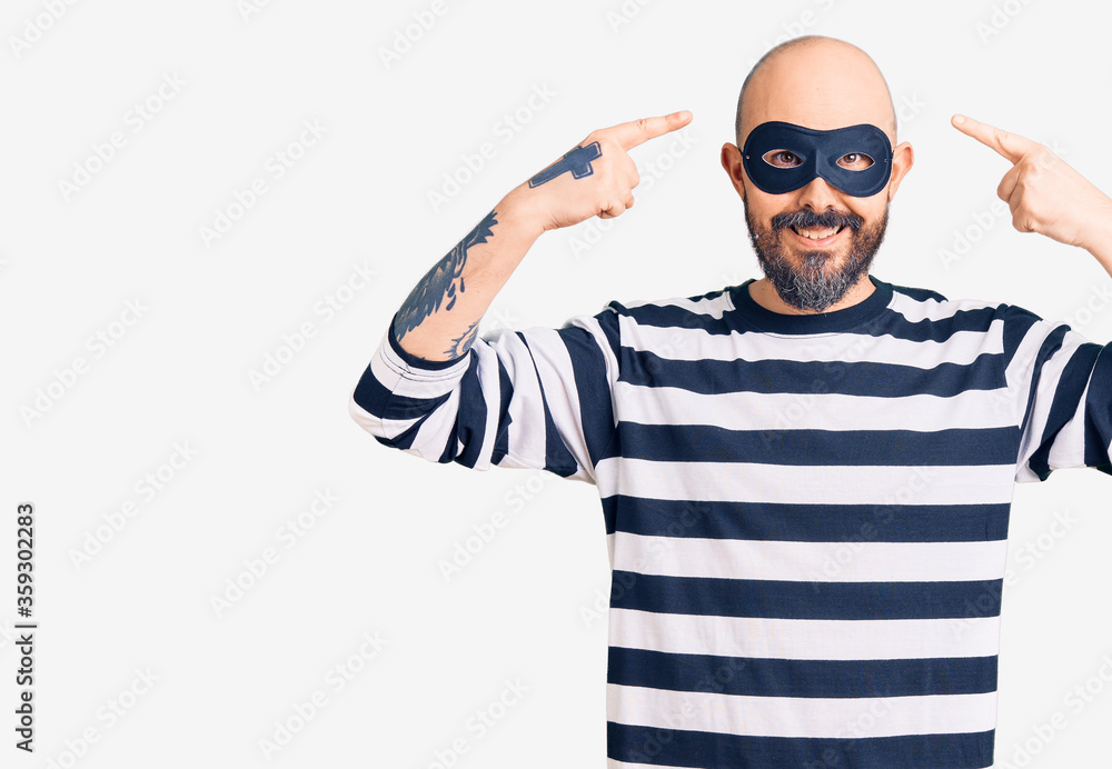 Young handsome man wearing burglar mask smiling pointing to head with both hands finger, great idea or thought, good memory Young handsome man wearing burglar mask smiling pointing to head with both hands finger, great idea or thought, good memory