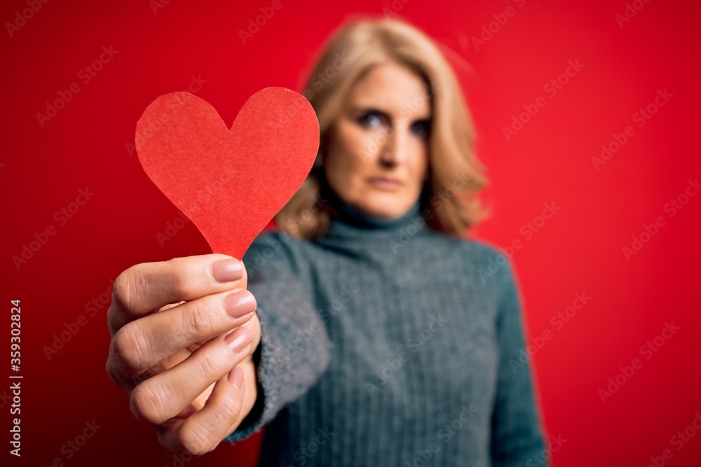 Middle age beautiful blonde romantic woman holding red paper heart celebrating valentine day with a confident expression on smart face thinking serious Middle age beautiful blonde romantic woman holding red paper heart celebrating valentine day with a confident expression on smart face thinking serious