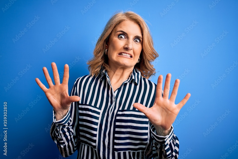 Middle age beautiful blonde woman wearing casual striped shirt standing over blue background afraid and terrified with fear expression stop gesture with hands, shouting in shock. Panic concept. Middle age beautiful blonde woman wearing casual striped shirt standing over blue background afraid and terrified with fear expression stop gesture with hands, shouting in shock. Panic concept.