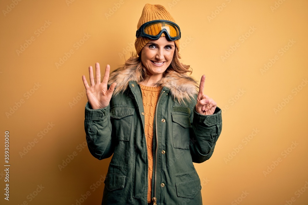 Middle age beautiful blonde skier woman wearing snow sportwear and ski goggles showing and pointing up with fingers number six while smiling confident and happy.