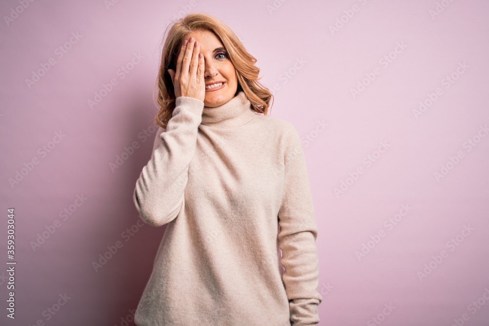 Middle age beautiful blonde woman wearing casual turtleneck sweater over pink background covering one eye with hand, confident smile on face and surprise emotion. Middle age beautiful blonde woman wearing casual turtleneck sweater over pink background covering one eye with hand, confident smile on face and surprise emotion.