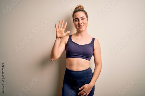 Young beautiful blonde sportswoman doing sport wearing sportswear over white background showing and pointing up with fingers number five while smiling confident and happy.