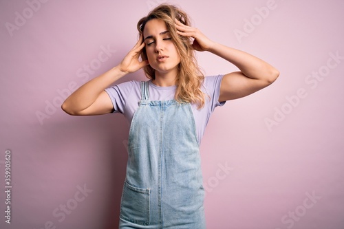 Young beautiful blonde woman wearing casual denim overalls standing over pink background with hand on head for pain in head because stress. Suffering migraine.