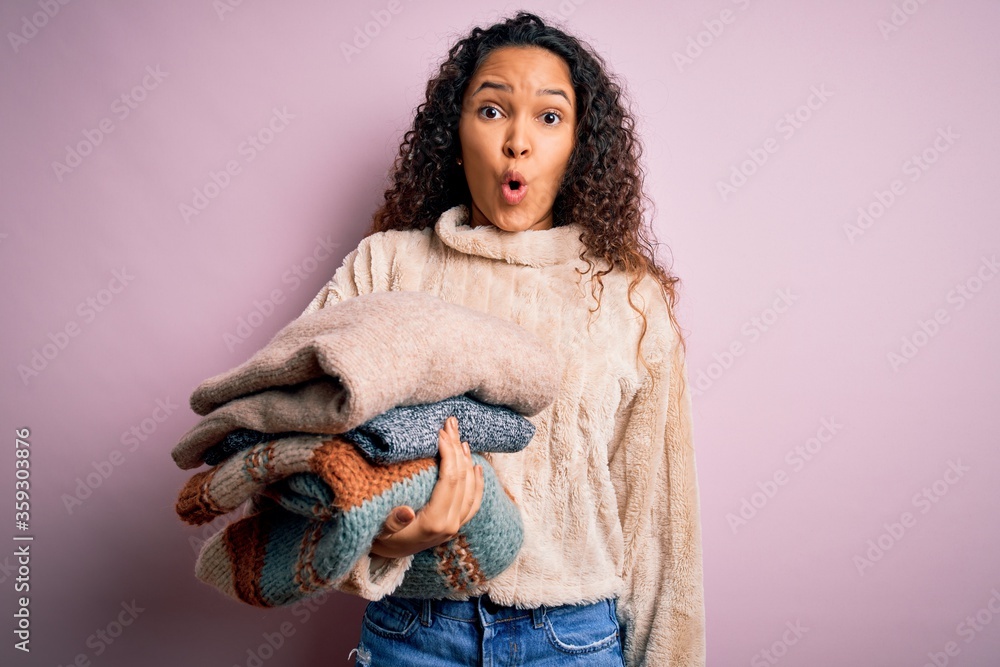 Beautiful shopkeeper woman with curly hair holding stack of clothes over pink background scared in shock with a surprise face, afraid and excited with fear expression Beautiful shopkeeper woman with curly hair holding stack of clothes over pink background scared in shock with a surprise face, afraid and excited with fear expression