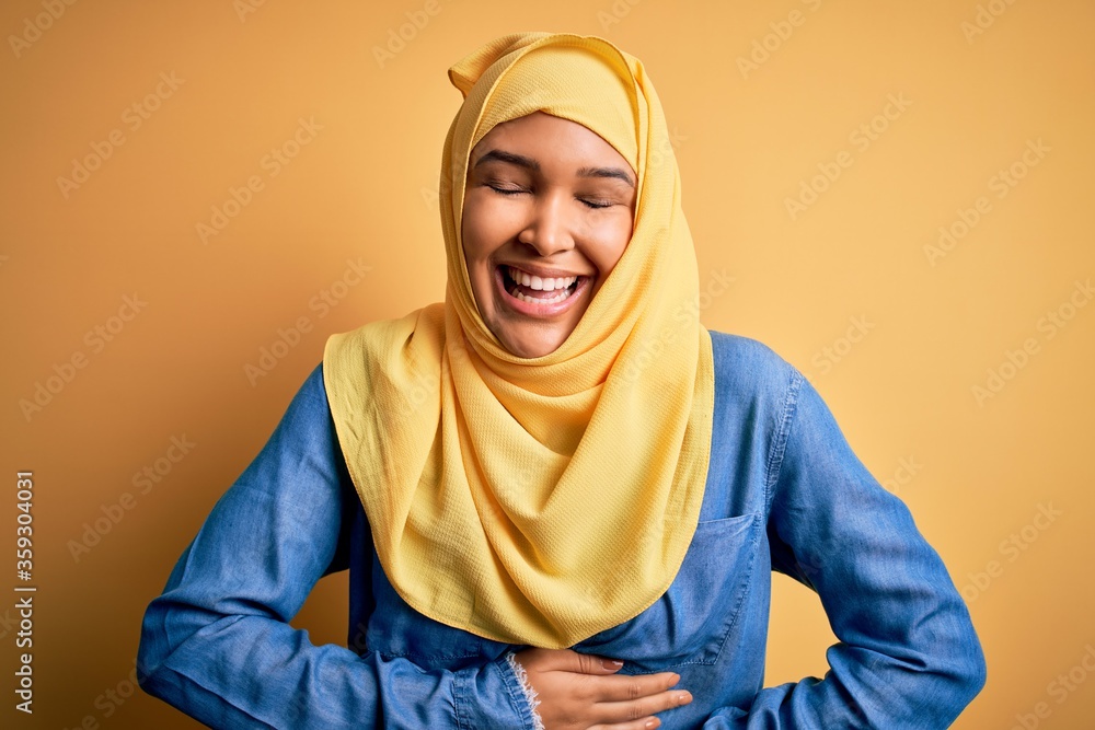 Young beautiful woman with curly hair wearing arab traditional hijab over yellow background smiling and laughing hard out loud because funny crazy joke with hands on body. Young beautiful woman with curly hair wearing arab traditional hijab over yellow background smiling and laughing hard out loud because funny crazy joke with hands on body.