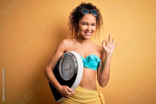 Young beautiful american slim woman on vacation wearing bikini holding weight machine doing ok sign with fingers  excellent symbol